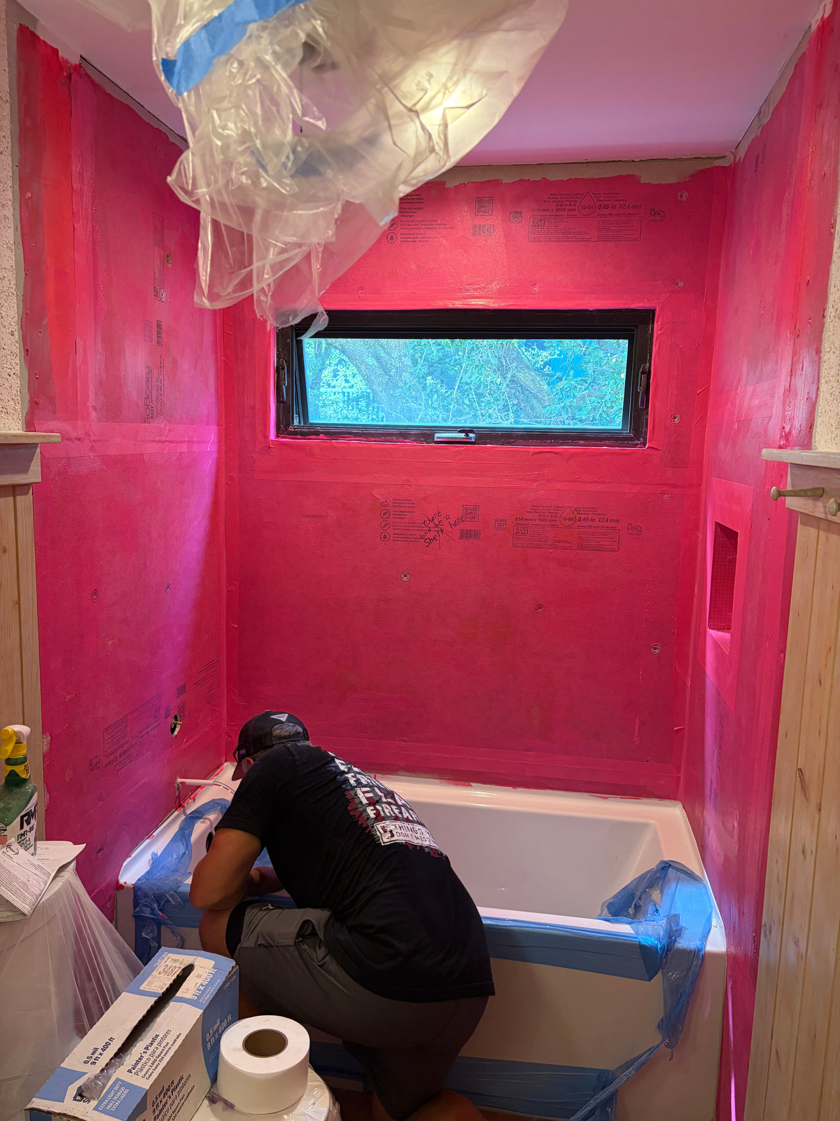 A person in a black t-shirt and gray shorts kneels beside a white bathtub in a bathroom under construction, with bright pink protective wall panels, blue painter's tape, plastic sheeting, and a small window visible.